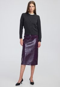 Vrouw in een donkergrijze geknoopte cardigan, glanzende donkerpaarse jurk tot de knie met een gestructureerd patroon, zwarte kitten heels, staand tegen een effen achtergrond.