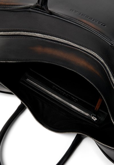 Intérieur d'un sac à main en cuir noir avec poche à fermeture éclair argentée, compartiment principal partiellement ouvert, et nom de marque embossé visible sur le rabat intérieur.