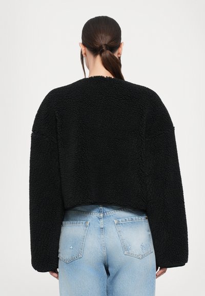 Veste noire courte en tissu texturé, avec des épaules tombantes et un design minimaliste. Associée à un jean bleu clair.