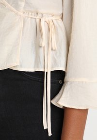 Ljusbeige blus med mjuk textur, som har en slinga i midjan och utsvängda ärmar, kombinerad med mörka denimjeans.