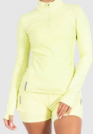 Kobieta w jasnożółtej sportowej bluzie na zamek z długim rękawem i pasujących szortach, stojąca na tle jednolitego, jasnego tła.