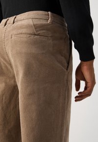 Vue latérale d'une personne portant un pantalon en velours côtelé beige et une chemise noire à manches longues, main détendue le long du corps.