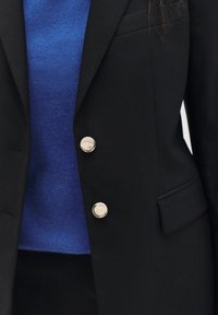 Blazer negro con botones plateados, usado sobre un suéter azul. El blazer presenta un diseño clásico y detalles en los bolsillos.