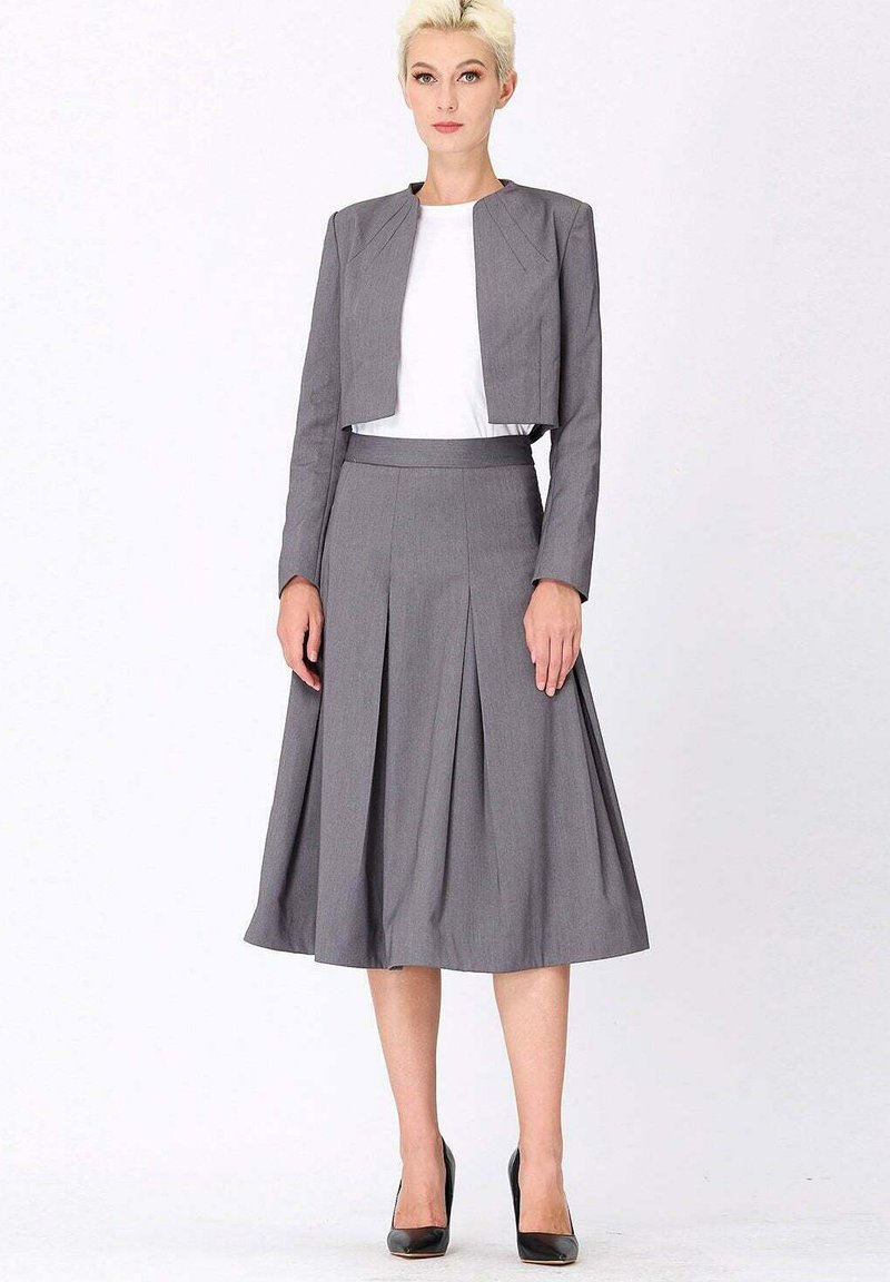 Femme portant un blazer court gris et une jupe midi plissée associée à un haut blanc et des talons hauts noirs, se tenant devant un fond blanc uni.