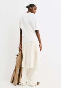 Witte oversized t-shirt, crèmekleurige rok met kantafwerking, beigekleurige suède jas over de arm, gecombineerd met lichte broek en zachte schoenen.