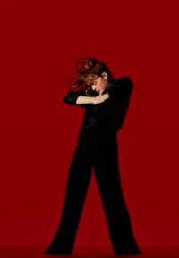 Abbigliamento tailored nero con maniche lunghe, in contrasto con uno sfondo rosso solido. I capelli sono lunghi e fluenti, catturati in movimento.