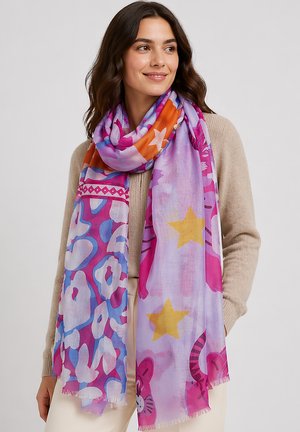 Frau trägt beigen Pullover und bunten Schal mit Sternen und abstrakten Mustern, lächelt und schaut zur Seite vor einfarbigem Hintergrund.