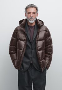 Brun pufferjakke med en blank finish og hætte, lagd over en mørkegrå blazer og en maroon skjorte. Jakken har lynlås.
