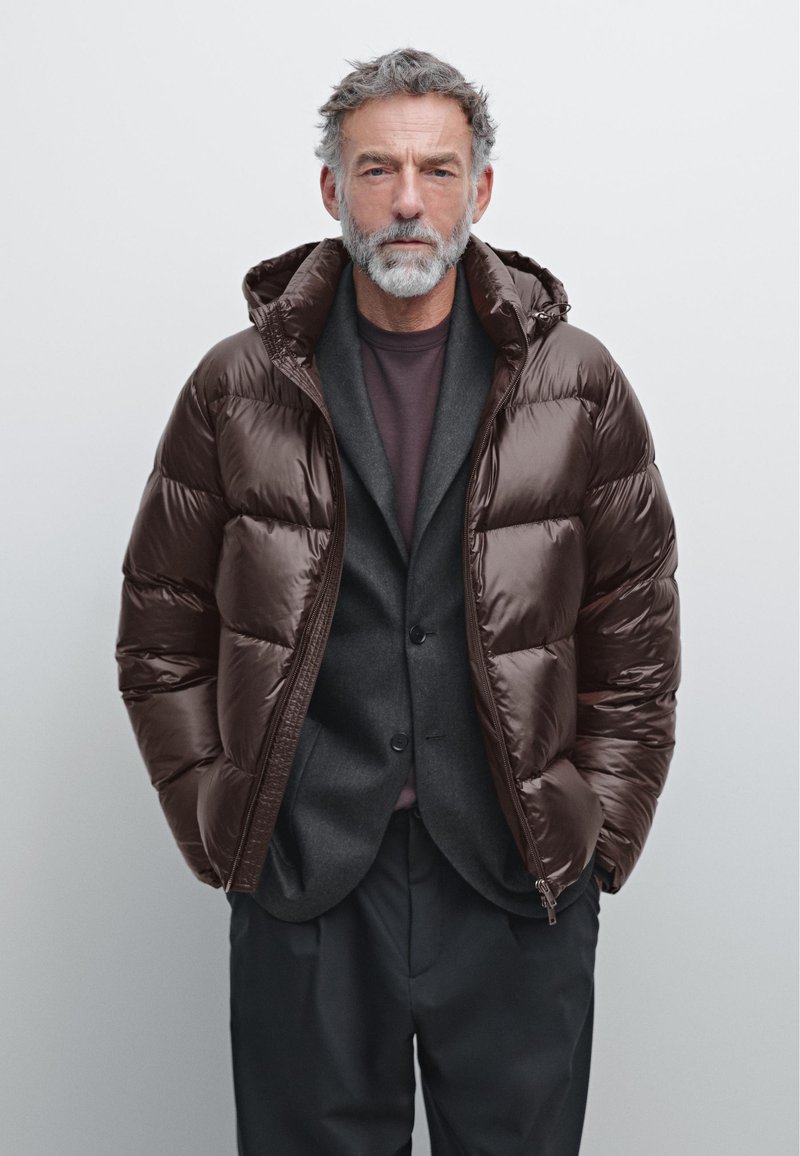 Brun pufferjakke med en blank finish og hætte, lagd over en mørkegrå blazer og en maroon skjorte. Jakken har lynlås.