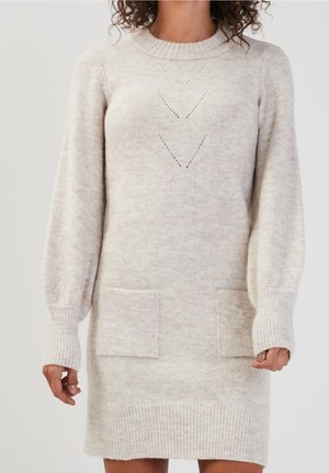 Femme portant une robe-pull beige en maille avec de longues manches bouffantes, des poignets côtelés, deux poches avant et des motifs en V subtils tricotés.