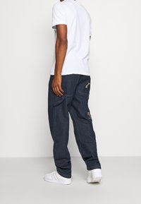 Hombre vistiendo una camiseta blanca, jeans cargo oscuros y holgados con parches, y zapatillas Adidas blancas, de pie contra un fondo blanco liso.