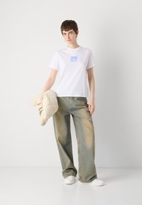 Tricou alb din bumbac cu un logo pătrat albastru, asortat cu o pereche de pantaloni din denim cu picioare largi, decolorati, și ținând în mână o jachetă puffer crem, cu matlasare.