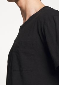 Černé bavlněné tričko s uvolněným střihem, výstřihem do V, krátkými rukávy a jednou kapsou na levé straně. Měkká textura a minimalistický design.