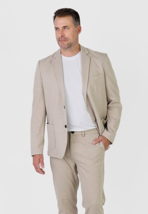 Mężczyzna w beżowym garniturze i białej koszuli, z ręką w kieszeni, spoglądający na bok na tle jednolitego jasnego tła.