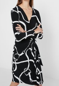 Svart klänning med djup V-hals, långa ärmar och ett vitt kedjemönster med klipp och rep. Mjuk tyg med en figursydd silhuett.