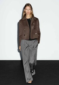 Brun dobbeltradet jakke lavet af uld, parret med grå plisserede bukser, der har en afslappet pasform og en glat tekstur.