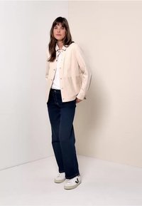 Lichtbeige button-up jack met textuurstof, gecombineerd met een wit shirt en donkerblauwe jeans. Accessoires omvatten witte sneakers.