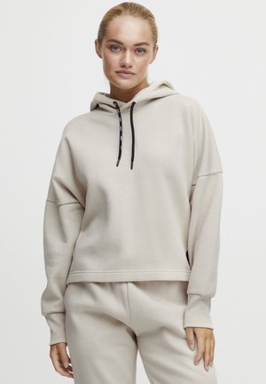 Beige hoodie with a loose fit, drawstrings, and ribbed cuffs. Smooth fabric texture and minimalist design. Worn with matching sweatpants.