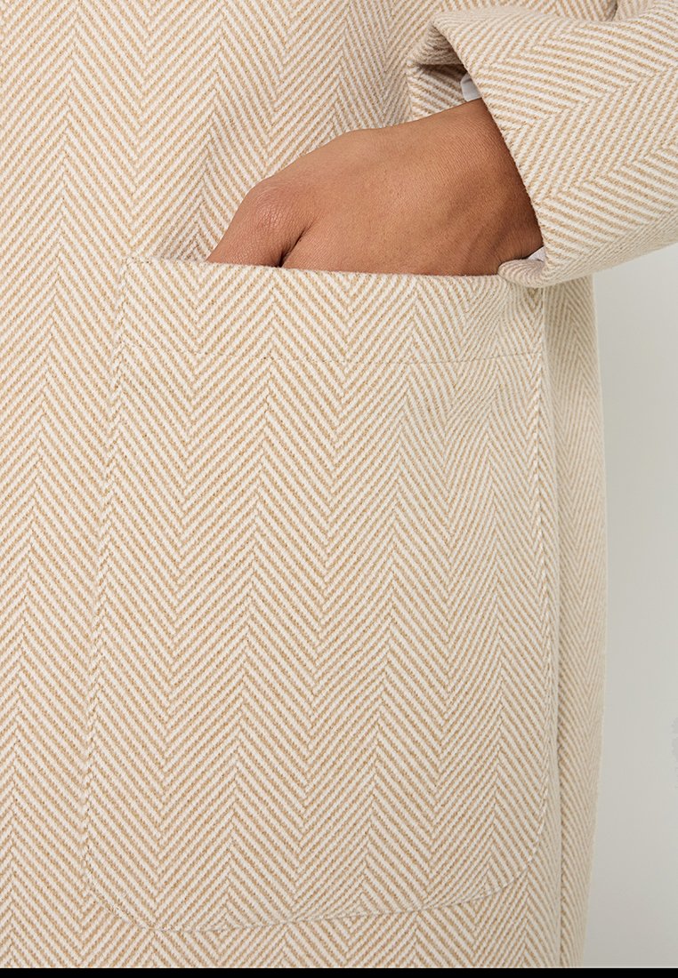 Beżowa tkanina w jodełkę o subtelnej teksturze. Obraz koncentruje się na kieszeni i dłoni, eksponując detale wzornictwa odzieży.