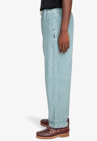 Light blue, loose-fitting pants made of soft fabric, featuring an elastic waistband and a side tag, paired with brown leather shoes.