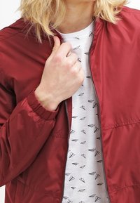Giubbotto bomber rosso in nylon con polsini elasticizzati, indossato sopra una camicia bianca con un motivo di aerei di carta neri. Dettaglio con zip sulla parte anteriore visibile.