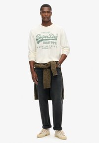 Crème lange mouw sweatshirt met groene print, gecombineerd met donkere jeans en beige sneakers. Bruin gestructureerd sweater om de taille geknoopt.