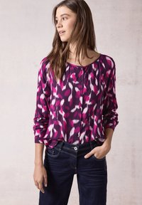 Blusa viola a maniche lunghe con motivo astratto rosa e bianco, scollatura rotonda e bottoni. Abbinata a jeans in denim scuro.