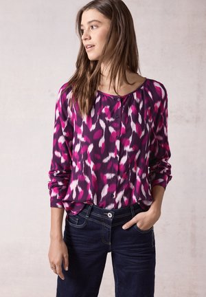 Blusa de manga larga de color púrpura con un patrón abstracto en rosa y blanco, escote redondo y tapeta de botones. Combinada con jeans de mezclilla oscura.
