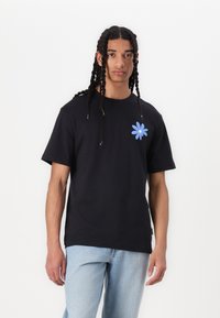 Sort bomulds t-shirt med rund halsudskæring. Har et lyseblåt blomstermotiv på venstre bryst. Bæres med lyseblå denimjeans.