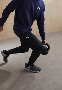 Námořnická mikina s logem, černé tepláky a šedé sportovní boty. Drží černou kettlebell. Hladká podlaha a neutrální pozadí.