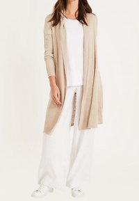 Beige lång cardigan i mjukt stickat material, öppen fram, kombinerad med en vit t-shirt och vida vita byxor, bärs med vita sneakers.