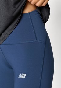 Tamno plave atletske tajice s glatkom teksturom, s tonalnim šavovima i bijelim New Balance logom na donjem dijelu noge.