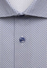 Gros plan d’une chemise à motifs bleus et blancs avec un col pointu et un seul bouton bleu fermé au centre.