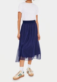 Sininen, hento, nilkkapituinen hame, jossa on kuminauhan vyötärö, pukeutuneena valkoisen t-paidan päälle ja yhdistettynä beigeihin ja vihreisiin lenkkareihin.