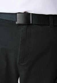 Ceinture en tissu noir avec une boucle rectangulaire mate portée sur un pantalon texturé foncé et une chemise blanche rentrée.