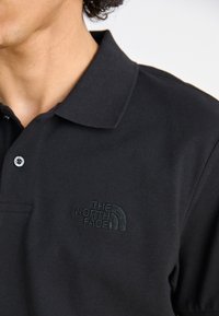Black polo shirt made from textured fabric, featuring a three-button placket and embossed logo on the left chest area.