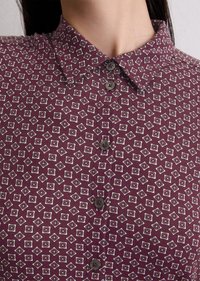 Chemise bordeaux avec un motif géométrique en blanc et rouge foncé, dotée d'un col boutonné et de quatre boutons foncés sur le devant.
