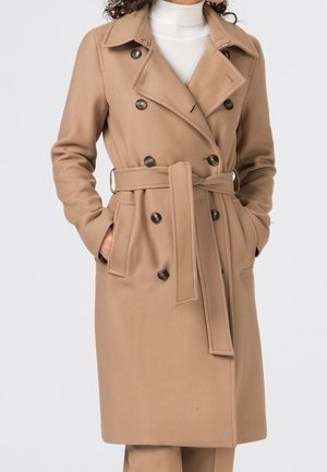 Manteau beige à double boutonnage, longueur genou, avec ceinture, fermetures boutons, poches latérales, porté sur un pull col roulé blanc.