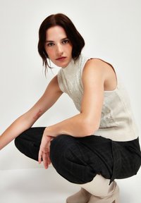 Top in maglia senza maniche di colore crema con motivo a trecce, abbinato a pantaloni palazzo a righe verticali scure e stivali alla caviglia beige. Modella accovacciata.