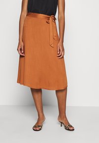 Woman wearing a rust-orange midi skirt with side tie, black sleeveless top, and leopard print open-toe mule heels, standing on gray floor.