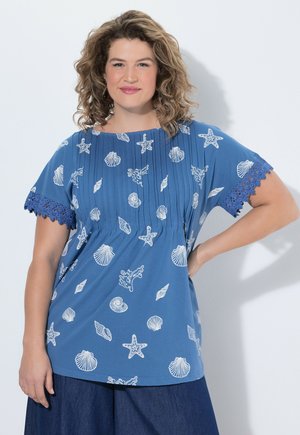 Kvinde med krøllet hår iført en blå top med hvide havtema-print og blonder på ærmerne, poserer med den ene hånd på hoften.