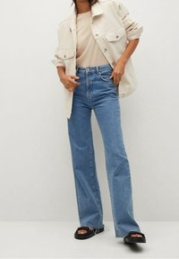 Denim flared jeans in light blue, featuring a high waist and five-pocket design. Worn with a beige top and cream oversized shirt. Black sandals.