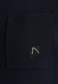 Maglione in tessuto strutturato blu navy con taschino quadrato sul petto; presenta un'etichetta con logo in bianco e nero sulla tasca.