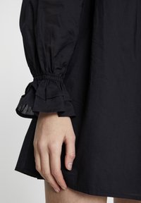 Robe noire avec manches bouffantes, poignets élastiqués froncés et détails à volants superposés, présentée près d'une main reposant sur le tissu.