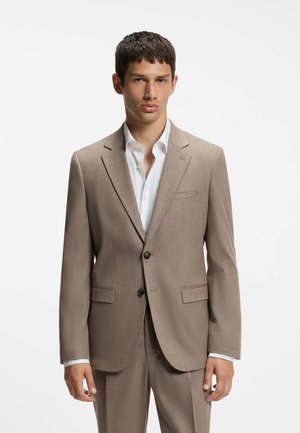 Joven vestido con un traje beige y camisa blanca de vestir, de pie frente a un fondo blanco liso.