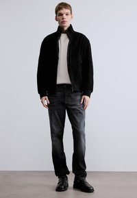 Fekete velúr dzseki krémszínű bordázott pulóver felett, sötét farmernadrággal és fekete cipővel. A modell egyenes testtartással áll egy minimalista háttér előtt.