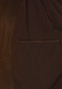 Primer plano de una tela de chaqueta marrón oscuro con un bolsillo con cremallera horizontal y un parche de cuero en el lado izquierdo.