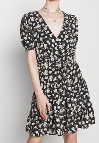 Robe noire à fleurs avec manches bouffantes, col en V et taille cintrée. Elle présente un ourlet volanté et de petits motifs de fleurs blanches et jaunes.