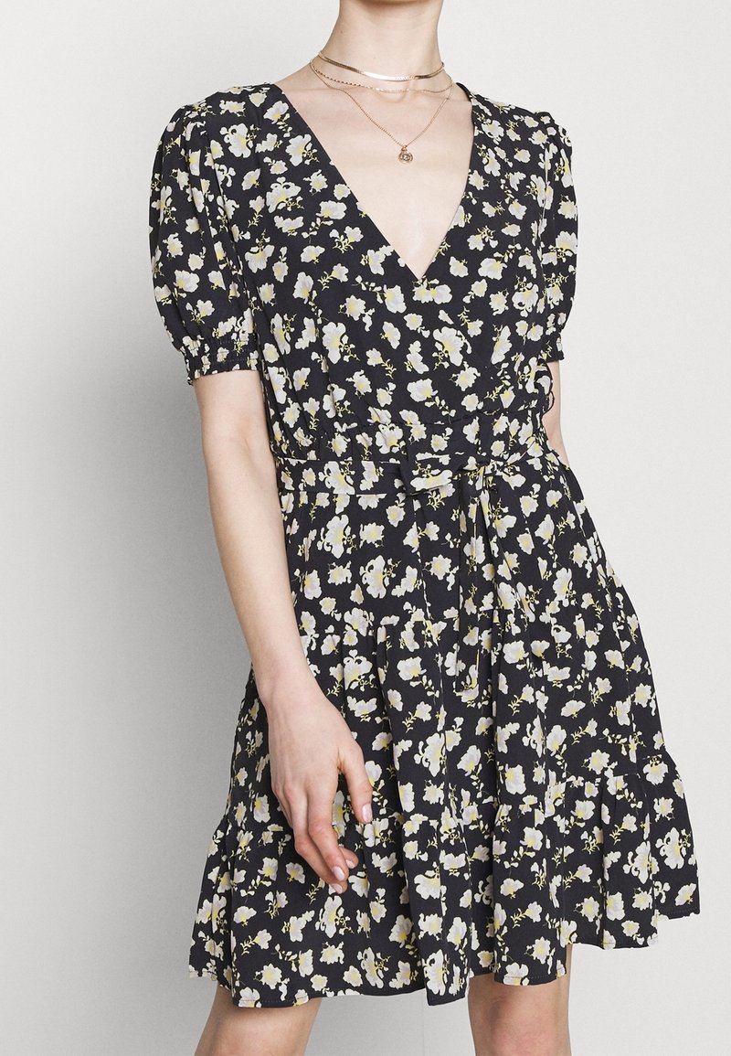 Robe noire à fleurs avec manches bouffantes, col en V et taille cintrée. Elle présente un ourlet volanté et de petits motifs de fleurs blanches et jaunes.