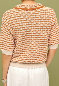 Gebreid sweater met een gestructureerd patroon van oranje en crèmekleurige zigzaglijnen, korte mouwen en een ribbelzoom, tentoongesteld tegen een groene achtergrond.